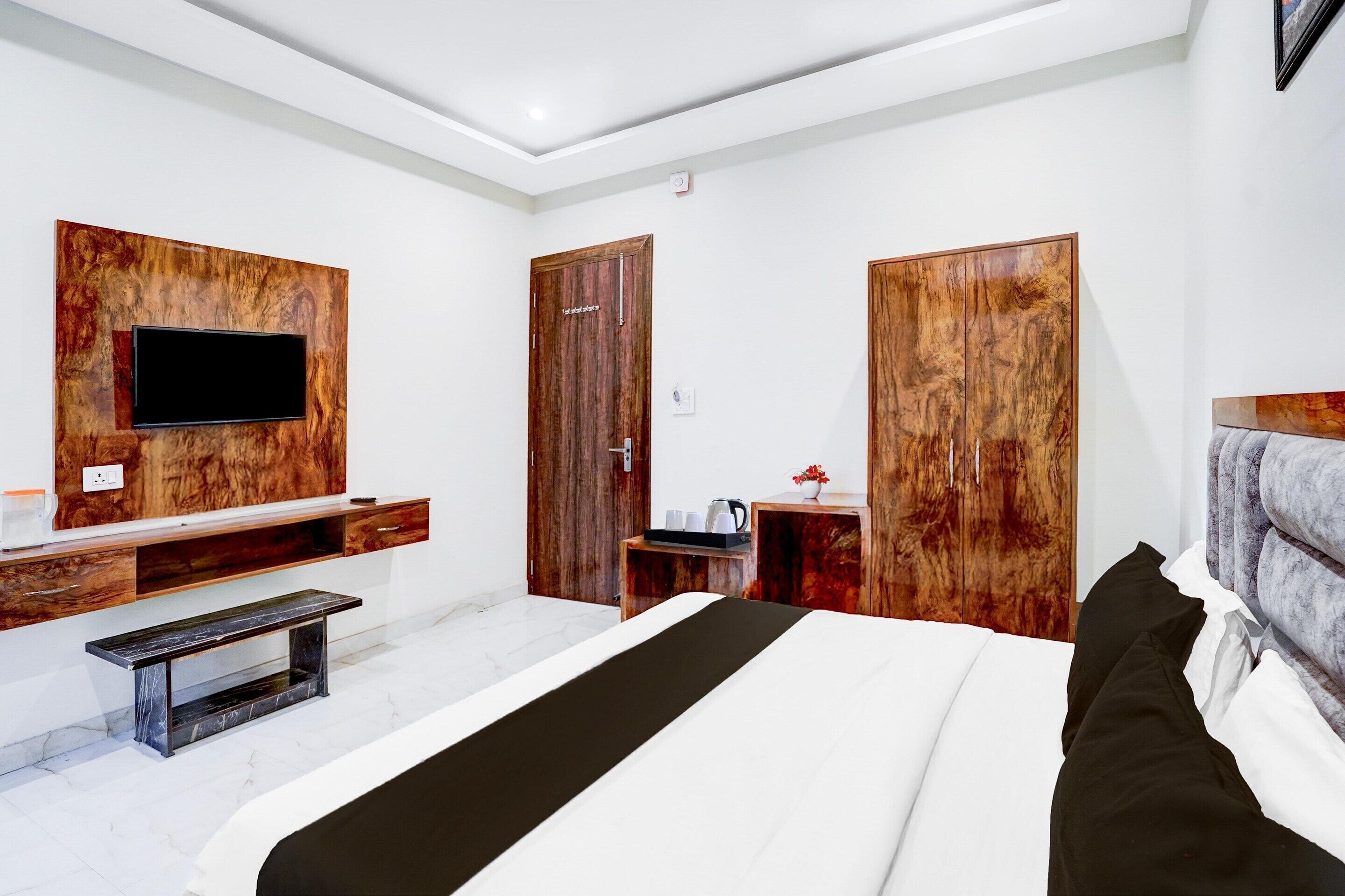 A modern hotel room with a large bed, a flat-screen TV mounted on a wooden panel, a wooden wardrobe, and a small table with a kettle.