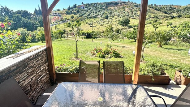 Een overdekt terras met een tafel en stoelen, met uitzicht op een groen landschap met bomen en heuvels.