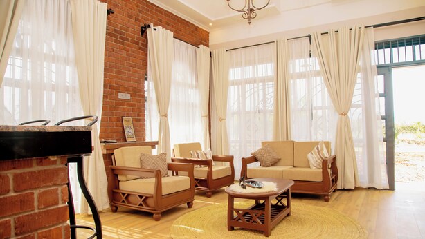 A living room with a brick wall, wooden furniture, and large windows with curtains. - Cosey Farm House and tent near  Ngorongoro, Serengeti and Manyara destinations. (Karatu - Arusha)