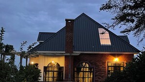 A two-story house with a dark roof, brick walls, and a car parked in front. - Cosey Farm House and tent near  Ngorongoro, Serengeti and Manyara destinations. (Karatu - Arusha)