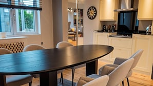 A modern dining area with a dark wooden table and light-colored chairs, a kitchen in the background, and a clock on the wall. - Stylish Dulwich Apartment With Parking (London)
