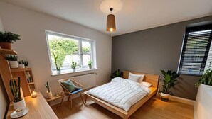 A modern bedroom with a wooden bed, a desk, and a chair. There are potted plants and a window with a view. - Stylish Dulwich Apartment With Parking (London)