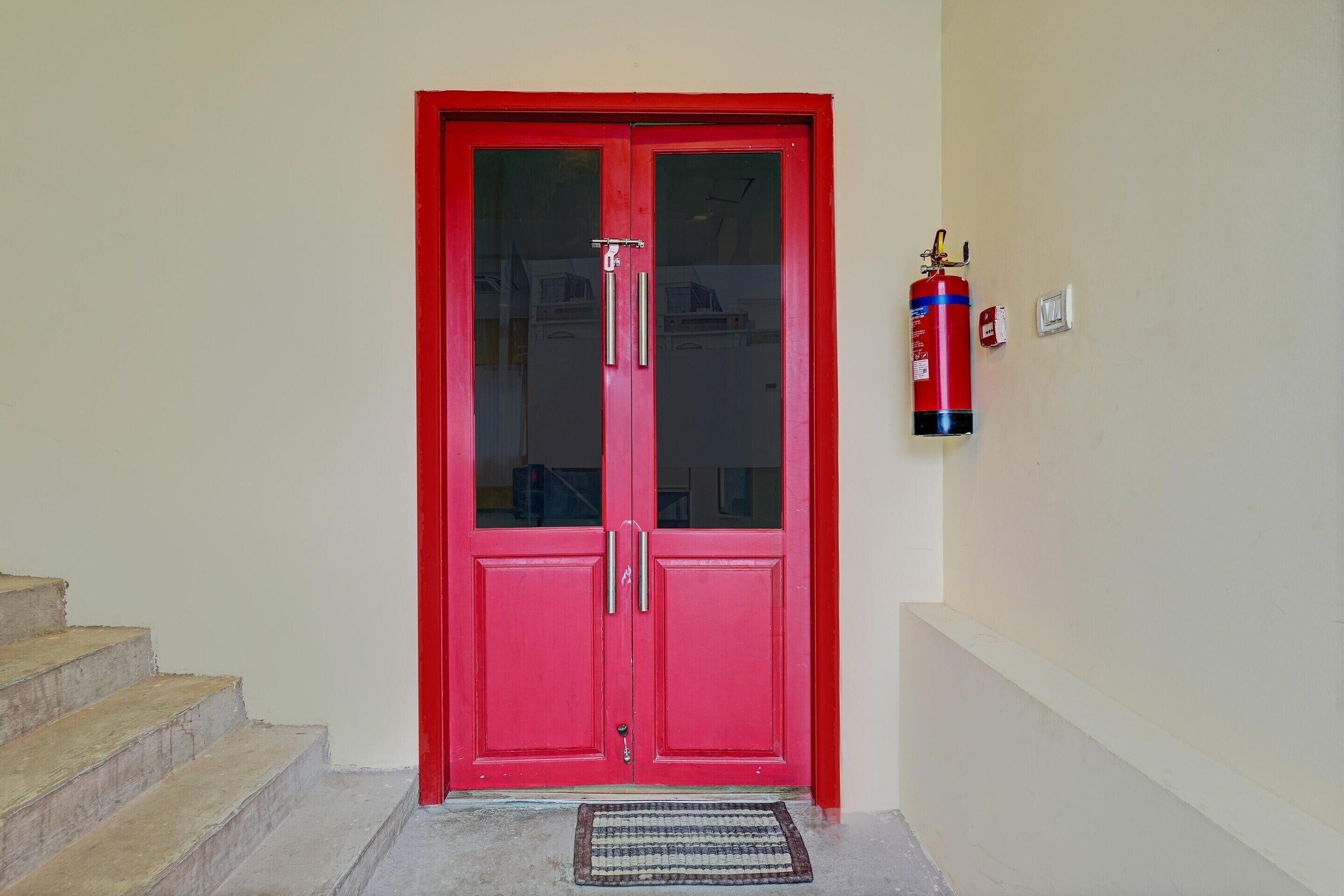 A red double door with glass panels, a fire extinguisher mounted on the wall to the right, and a set of concrete steps leading up to the door.