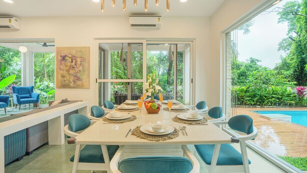 A dining area with a table set for six, surrounded by chairs, with a view of a pool and greenery outside. - ELIVAAS Casa Del Mar 4BHK Villa & Pool (Anjuna)