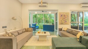 A modern living room with a sofa, a coffee table, and a TV. There is a window with a view of greenery outside. - ELIVAAS Casa Del Mar 4BHK Villa & Pool (Anjuna)
