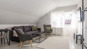 A modern loft apartment with a high ceiling, a gray sofa, a striped armchair, a small table, and a large window with sheer curtains.