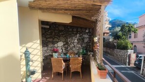 Un balcón con sillones de mimbre, una mesa, un muro de piedra y vistas a una calle y edificios.