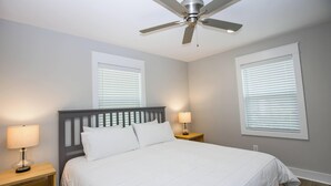 Ein Schlafzimmer mit einem Bett, Nachttischen, Lampen, einem Deckenventilator und zwei Fenstern mit Jalousien.