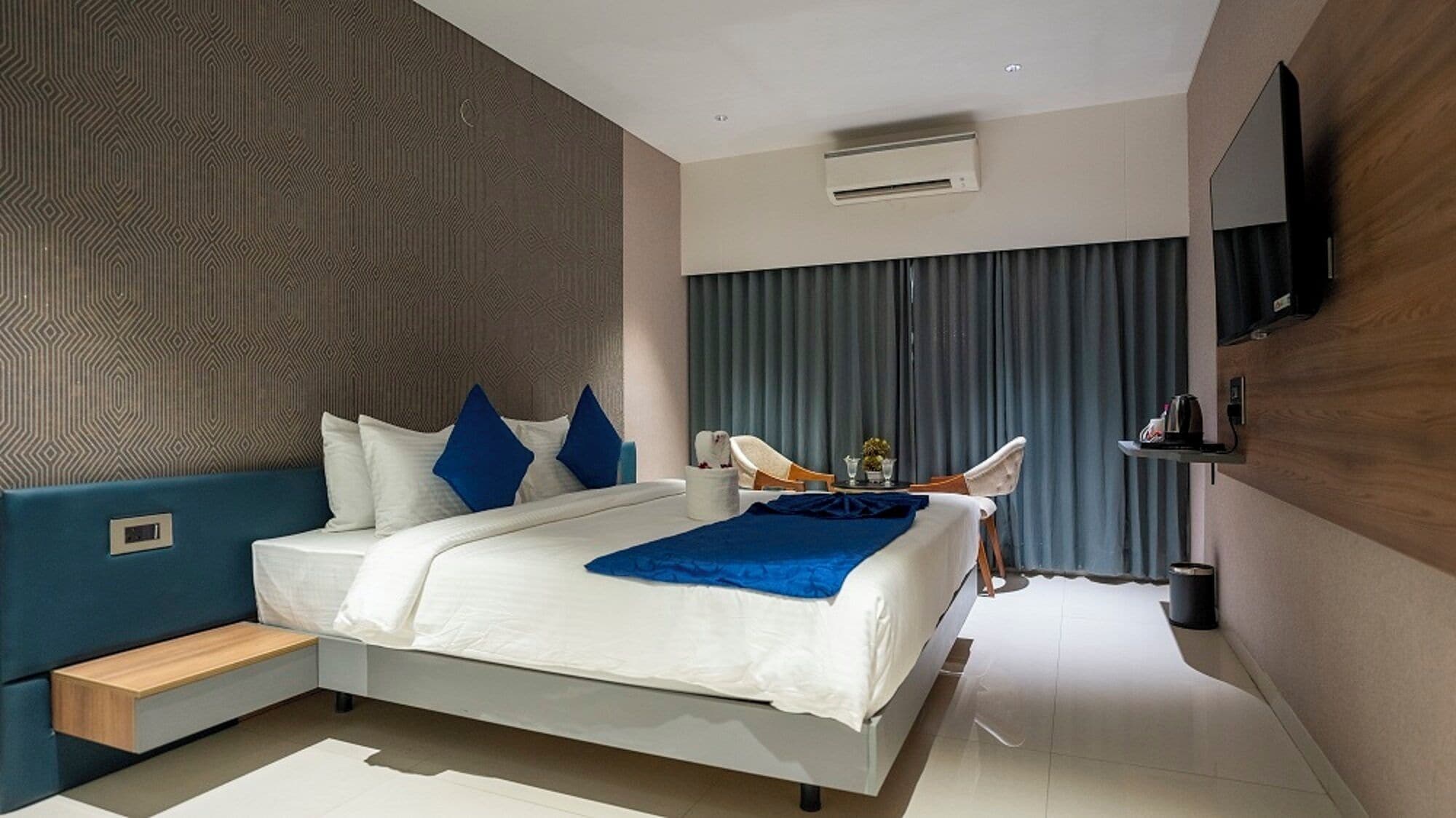 A modern hotel room with a bed, a seating area, a TV, and an air conditioning unit.