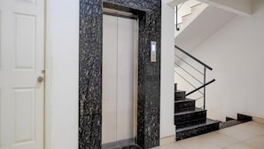 A modern elevator with a granite facade, a white door, and a staircase with black steps and metal railings. - Deluxehotel Gurudev Residency (Bengaluru)