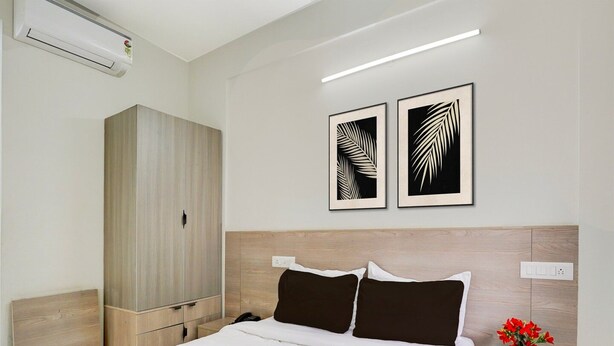 A hotel room with a bed, a wardrobe, two wall-mounted artworks, a bedside table with a vase of flowers, and a modern light fixture. - Deluxehotel Gurudev Residency (Bengaluru)