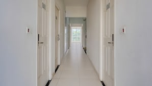 A long, narrow hallway with white walls and doors, leading to a bright room with a window. - Deluxehotel Gurudev Residency (Bengaluru)