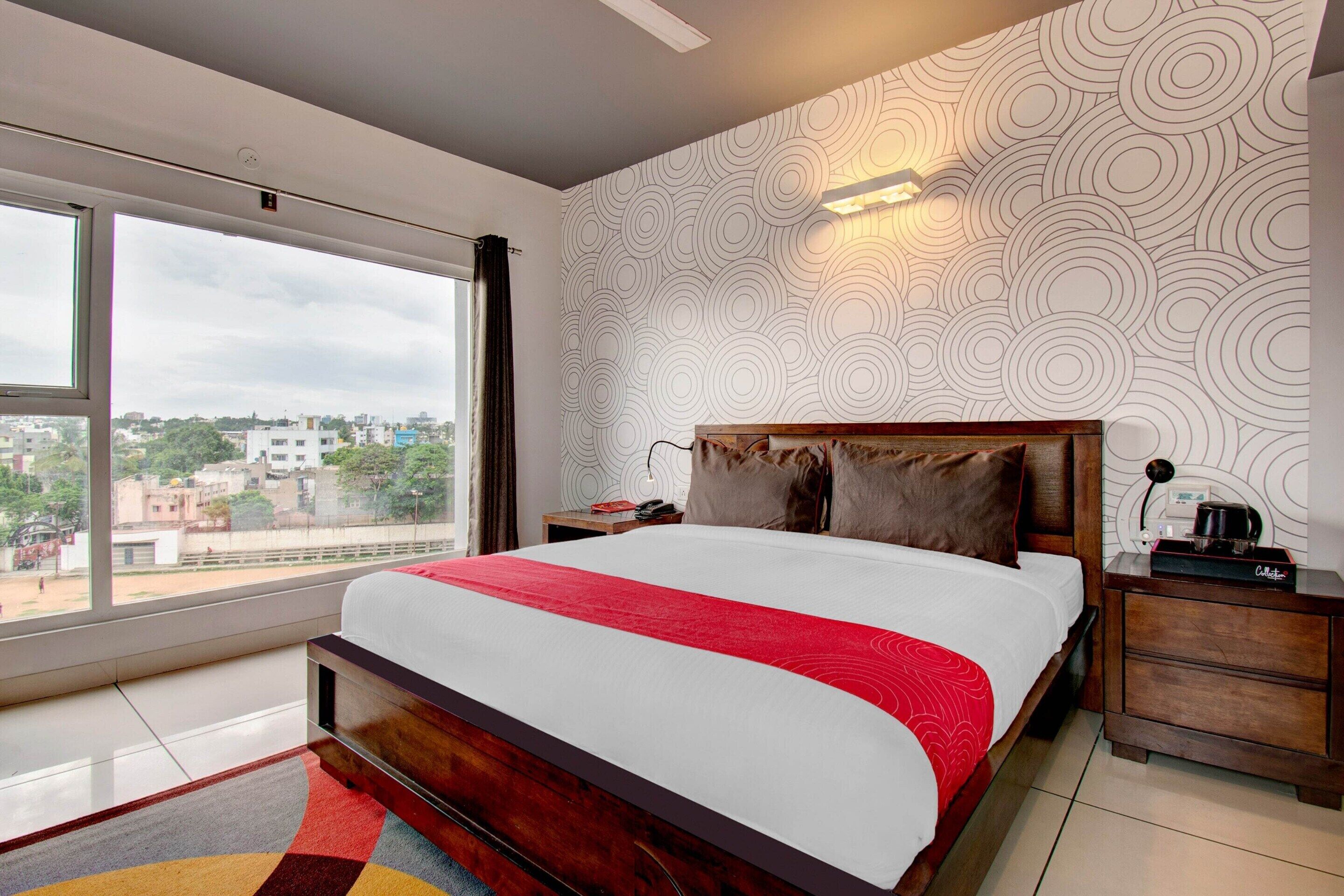 Une chambre d’hôtel avec une grande fenêtre, un lit recouvert d’une couverture rouge, une tête de lit en bois, une table de chevet avec une lampe et un mur orné d’un motif.