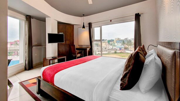 A hotel room with a large bed, a desk, a chair, a TV, and a view of the city. - Deluxehotel Palm Tree (Bengaluru)