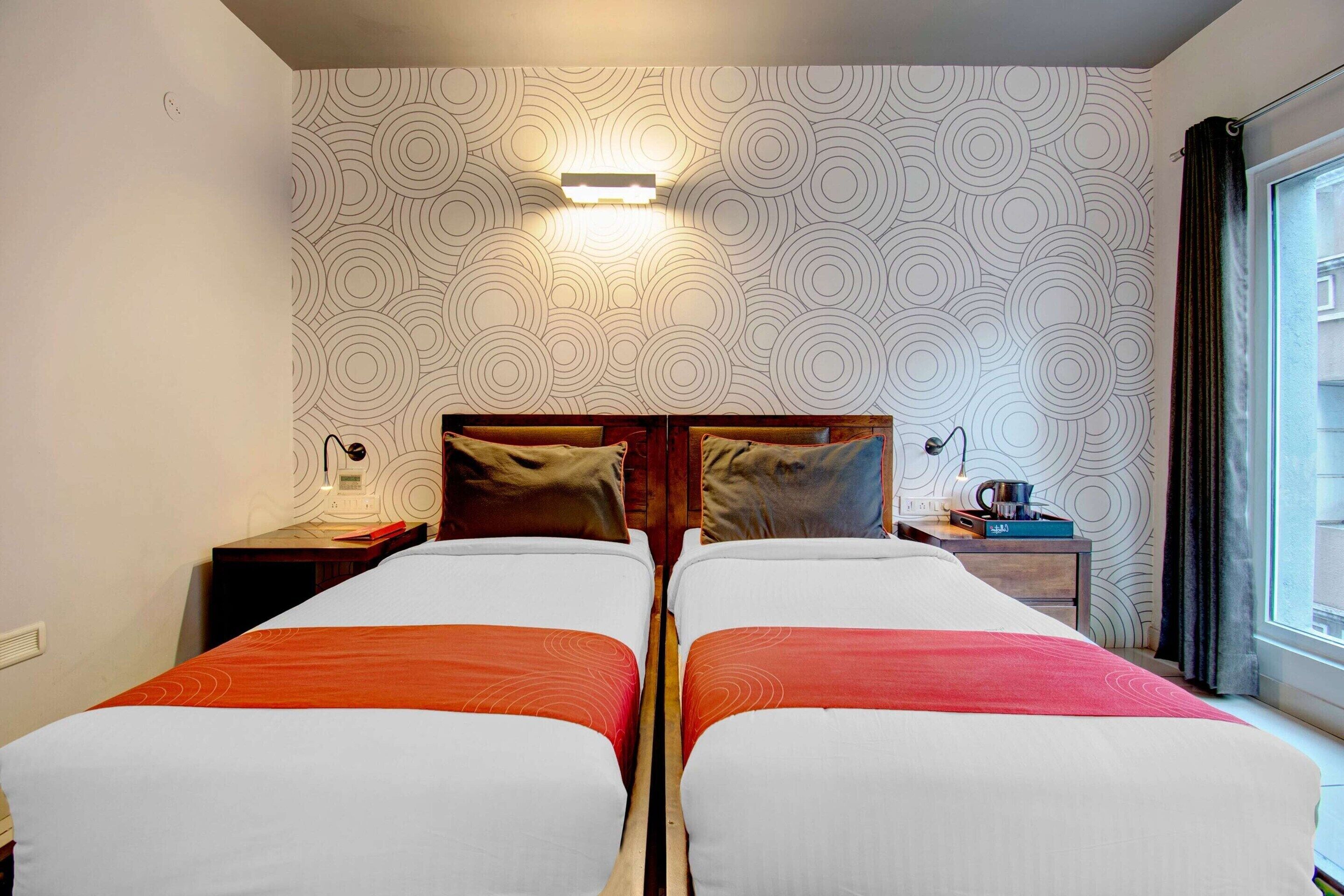 Habitación de hotel con dos camas, una mesita de noche con lámpara, una pequeña mesa con hervidor y una ventana con cortinas.
