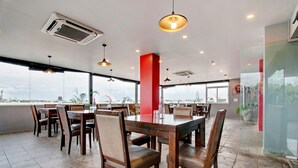 A modern dining area with wooden tables and chairs, large windows offering a view, and pendant lighting. - Deluxehotel Palm Tree (Bengaluru)