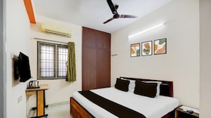 Une chambre à coucher comprenant un lit, un ventilateur de plafond, un climatiseur, une fenêtre avec des rideaux, une petite table, un téléviseur et des œuvres d’art encadrées accrochées au mur.
