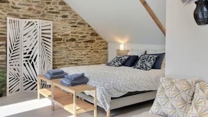 Un dormitorio con techo inclinado, una cama con ropa de cama estampada, una mesita de noche con toallas, una planta colgante en la pared y un panel decorativo.