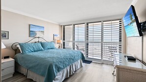 Ein Schlafzimmer mit Bett, Nachttisch, Schreibtisch, Fernseher und einem Fenster mit Jalousien.