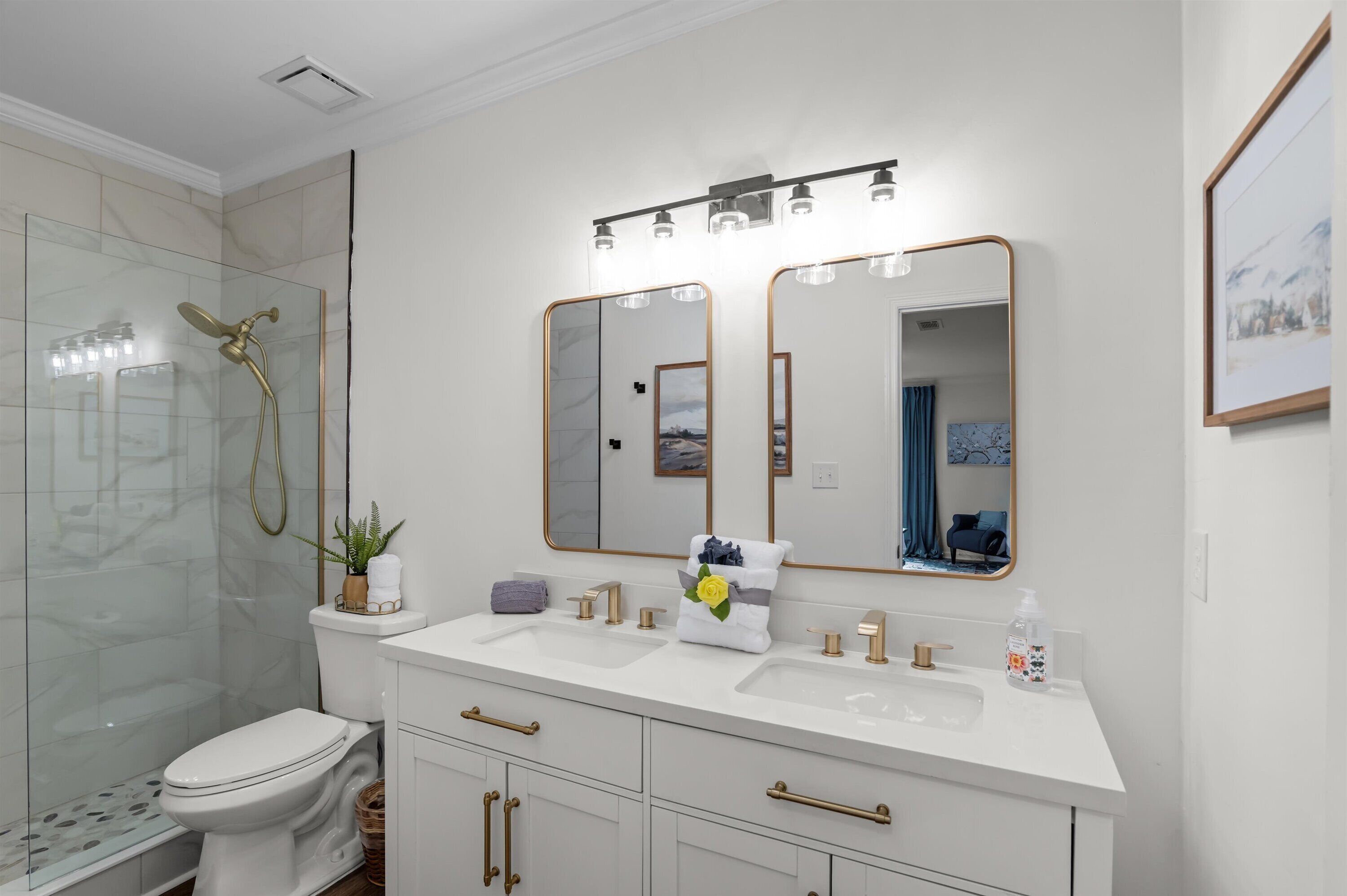 Une salle de bain moderne dotée d’une douche à parois de verre, de toilettes, d’un meuble-lavabo avec deux vasques et d’un grand miroir avec un cadre doré.