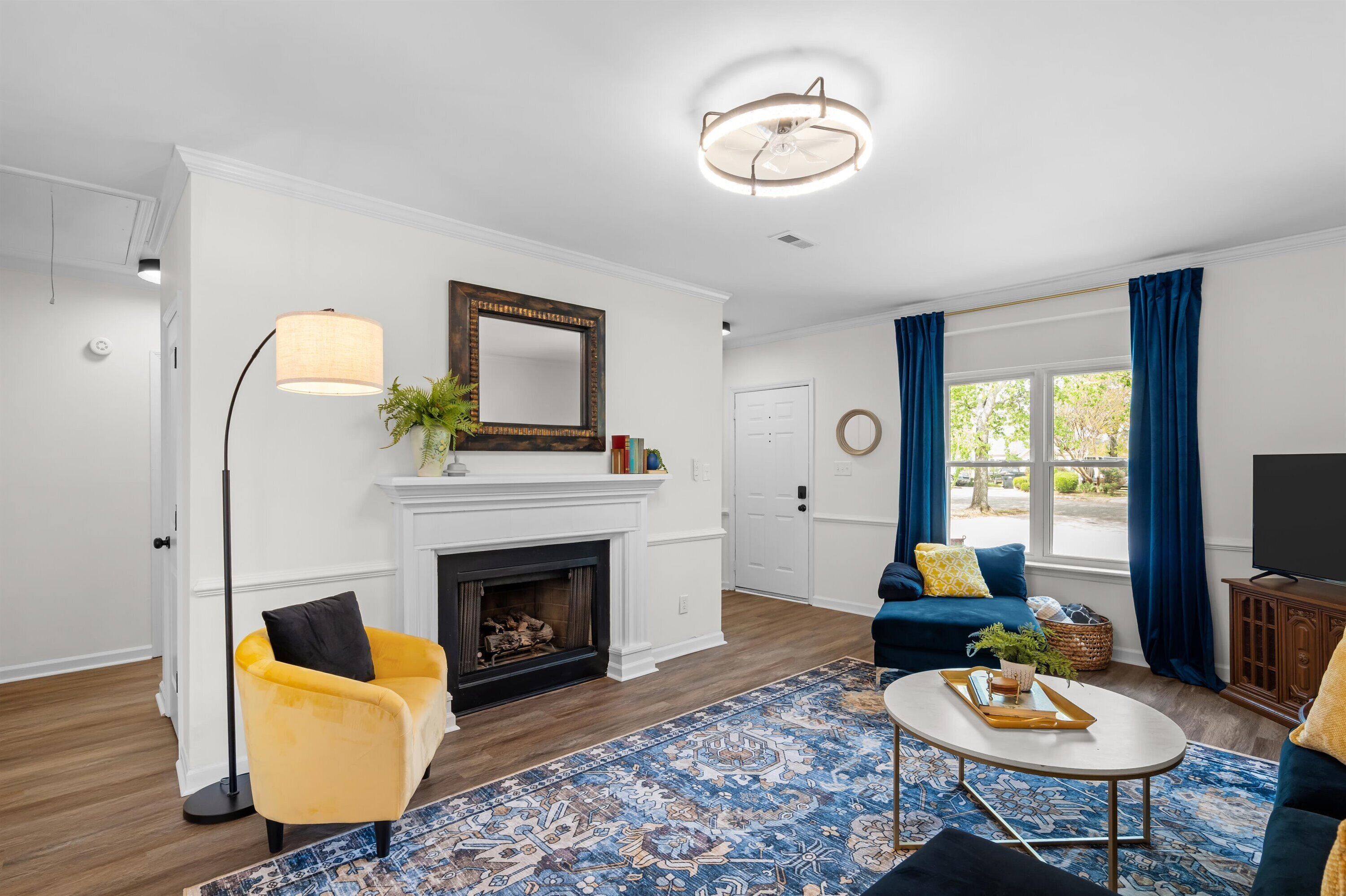 Una sala de estar con chimenea, un sofá, un sillón amarillo, una mesa de centro y un ventanal con cortinas.