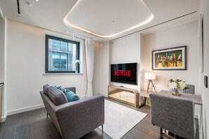 A modern living room with a sofa, a TV displaying 'NETFLIX,' a dining area with a table and chairs, and a window with curtains. - Luxury Residence - TCRW Soho Square (London)