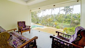 A room with a large glass door leading to a pool and palm trees, wooden furniture, and a view of the outdoors. - Deluxe Thiruvallam Lakeview (Thiruvananthapuram)