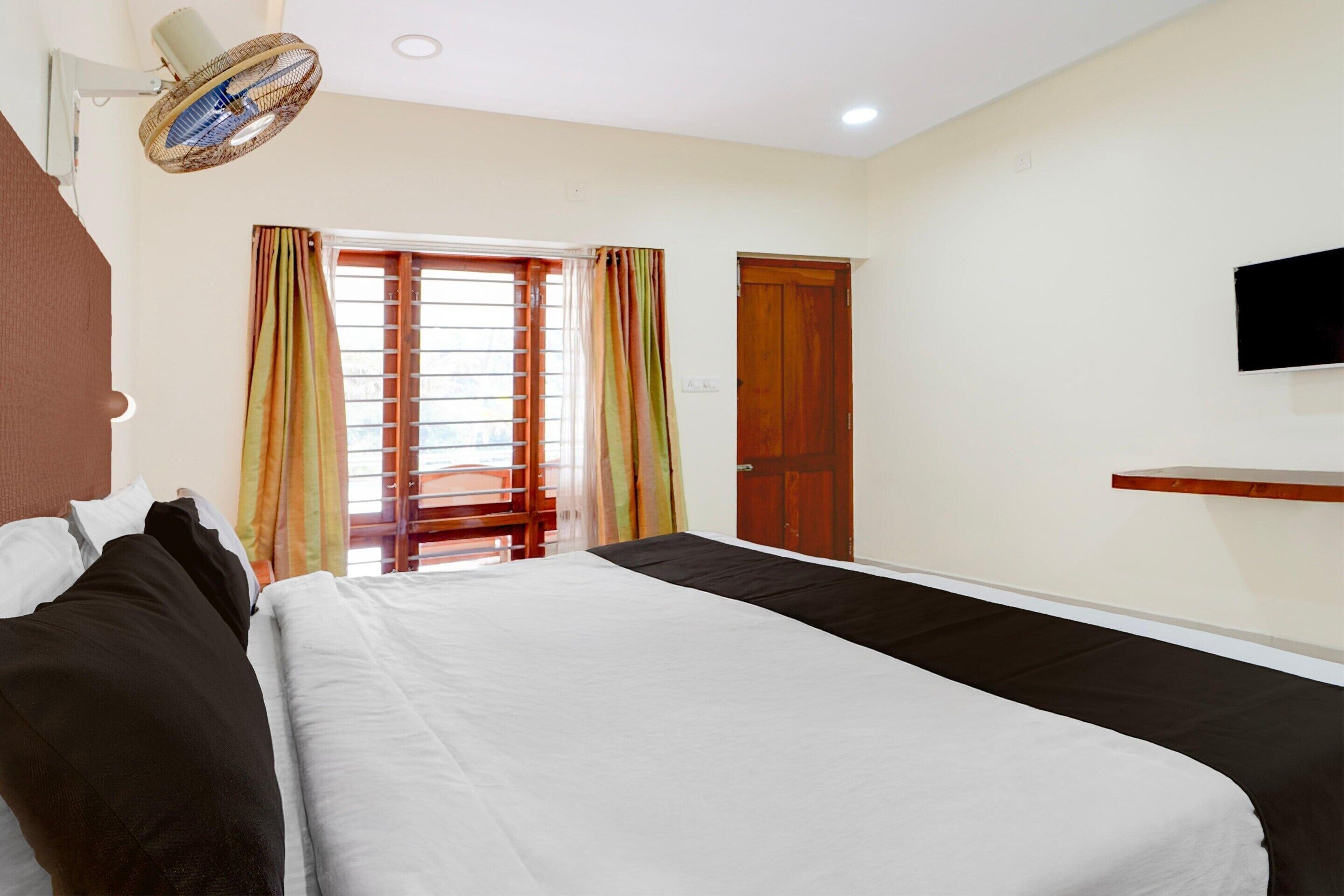 A hotel room with a bed, a TV mounted on the wall, a window with curtains, and a ceiling fan.