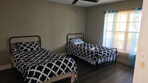 Dos llits individuals amb roba de llit estampada en una habitació amb ventilador de sostre, una finestra amb cortines i un terra de fusta.