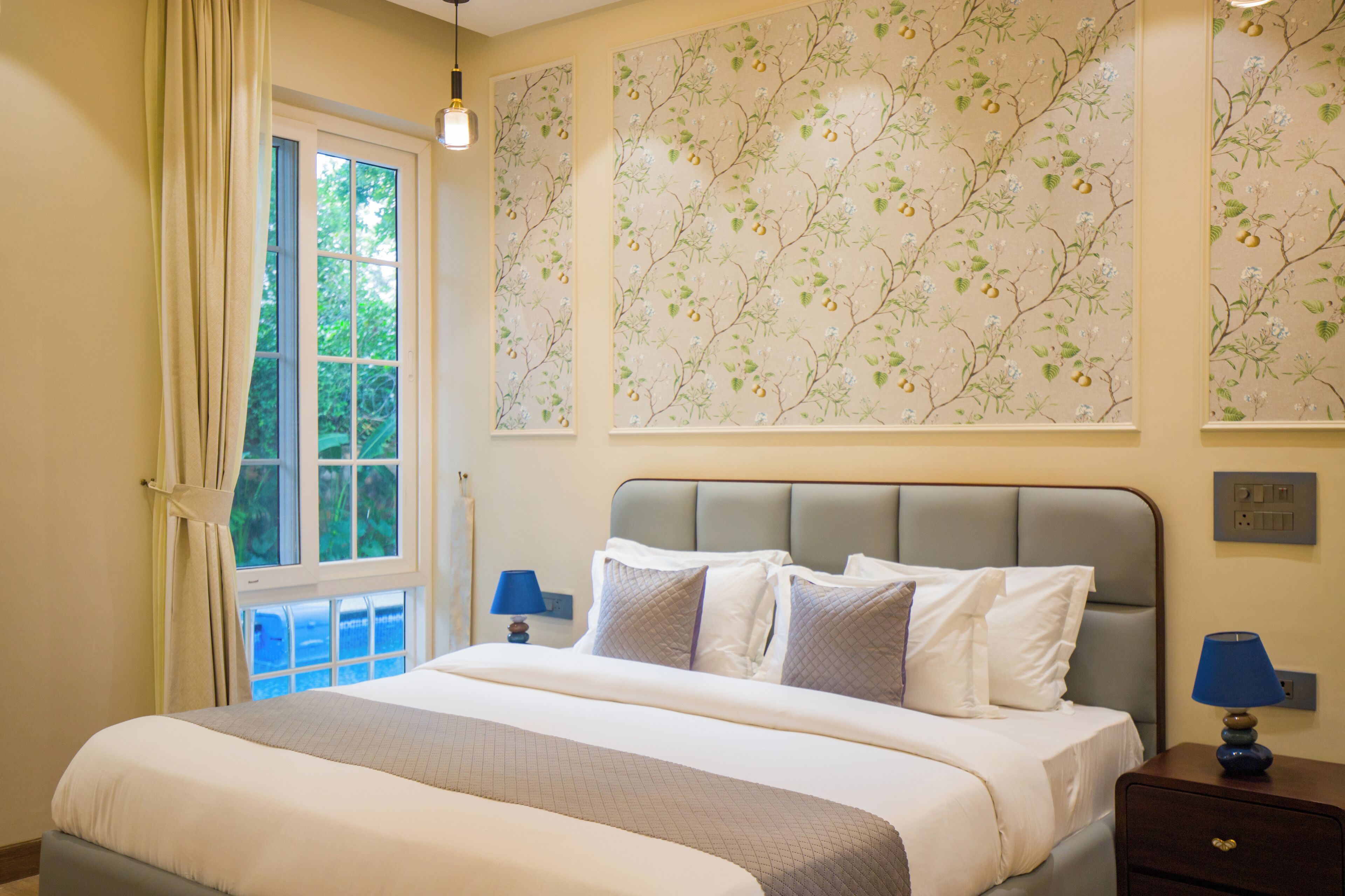 Een slaapkamer met een bed, nachtkastje, lamp, raam met gordijnen en een bloemen behang.