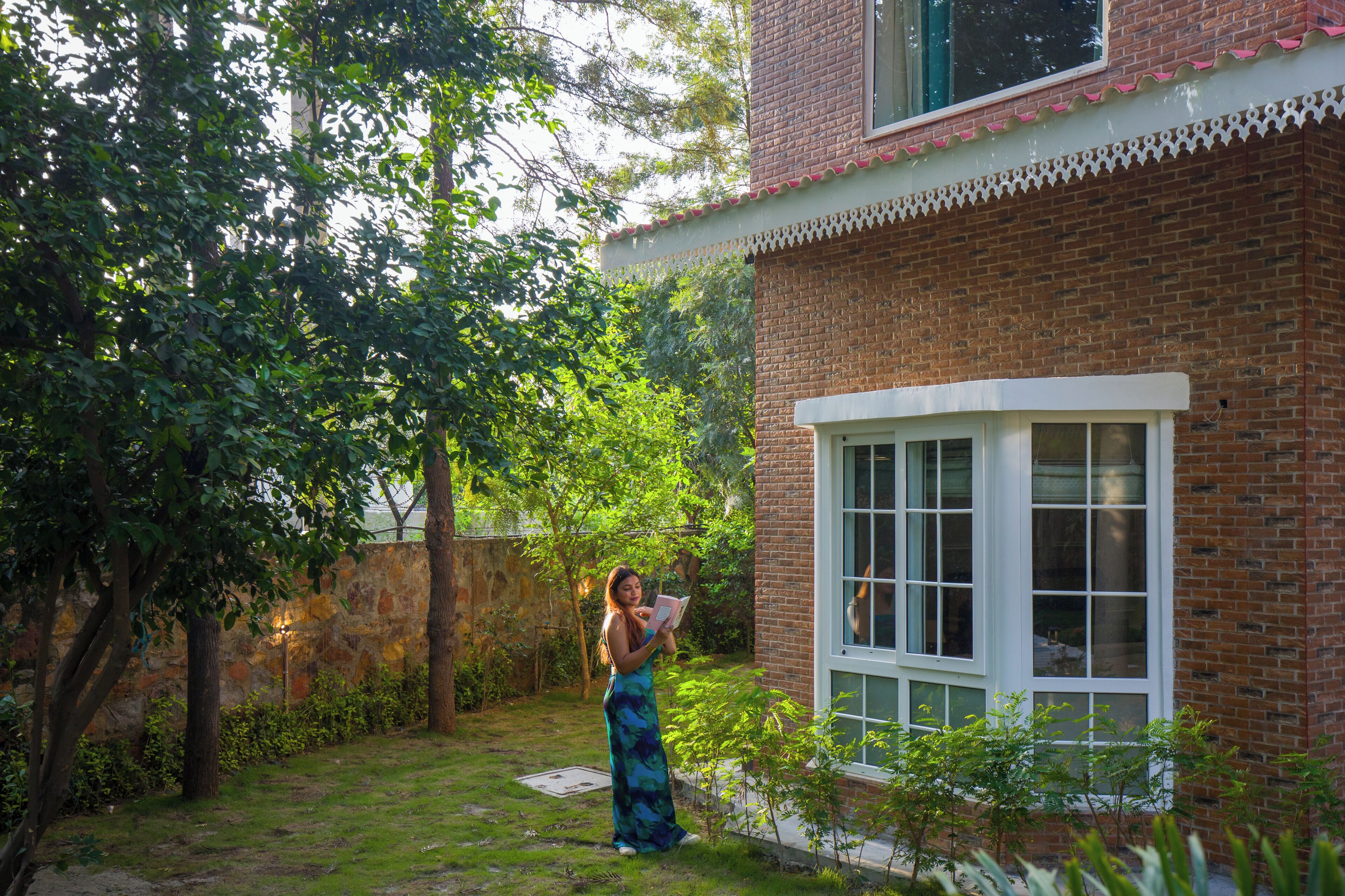 A brick building with a white window, a stone wall, and a garden with trees and grass.