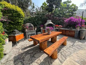 Une terrasse en pierre avec une table et des bancs en bois, entourée de plantes en pot et d’un four en pierre.