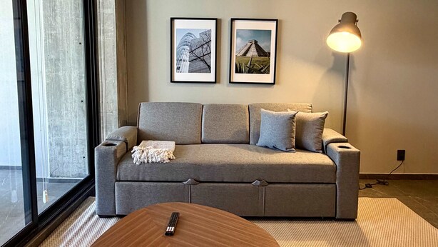 Ein modernes Wohnzimmer mit einem grauen Sofa, zwei gerahmten Bildern an der Wand, einer Stehlampe und einer Fernbedienung auf einem runden Tisch.