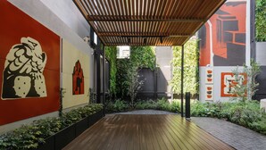 Eine Holzterrasse führt zu einem überdachten Bereich mit moderner Architektur, darunter eine Pergola und eine Wand mit abstrakter Kunst.