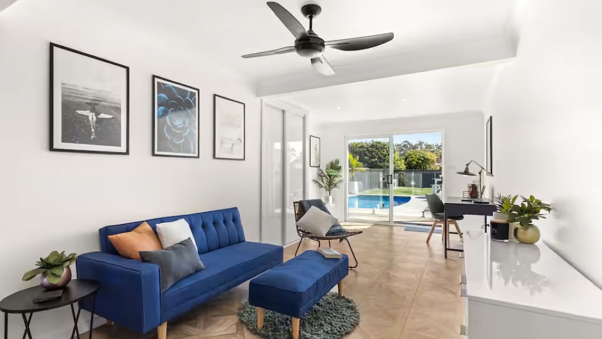 Ein modernes Wohnzimmer mit einem blauen Sofa, einem Deckenventilator und Blick auf einen Swimmingpool.