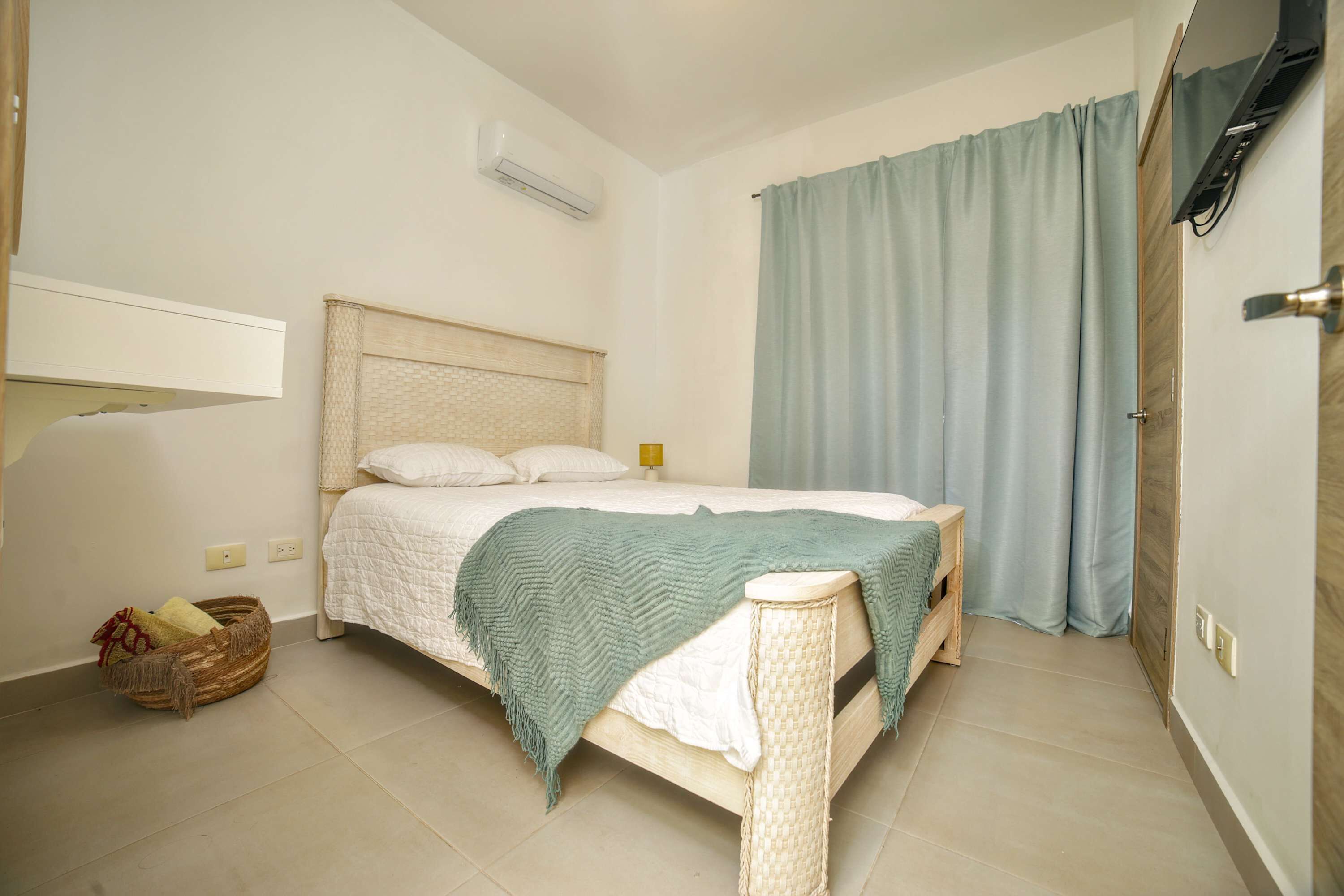 A bedroom with a bed, a bedside table, a wall-mounted TV, and a window with curtains.