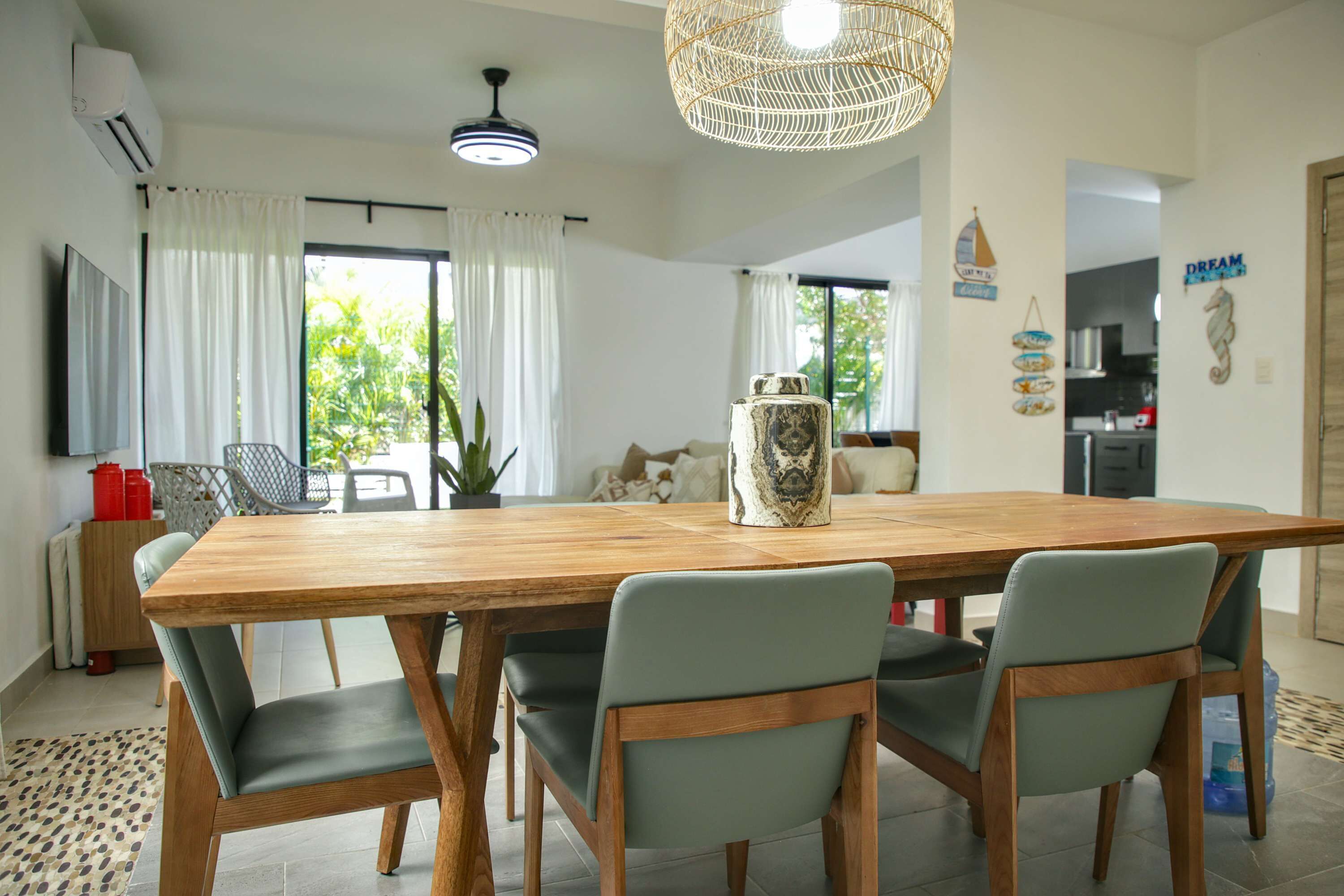 A dining area with a wooden table, four chairs, a hanging light fixture, and a kitchen in the background.
