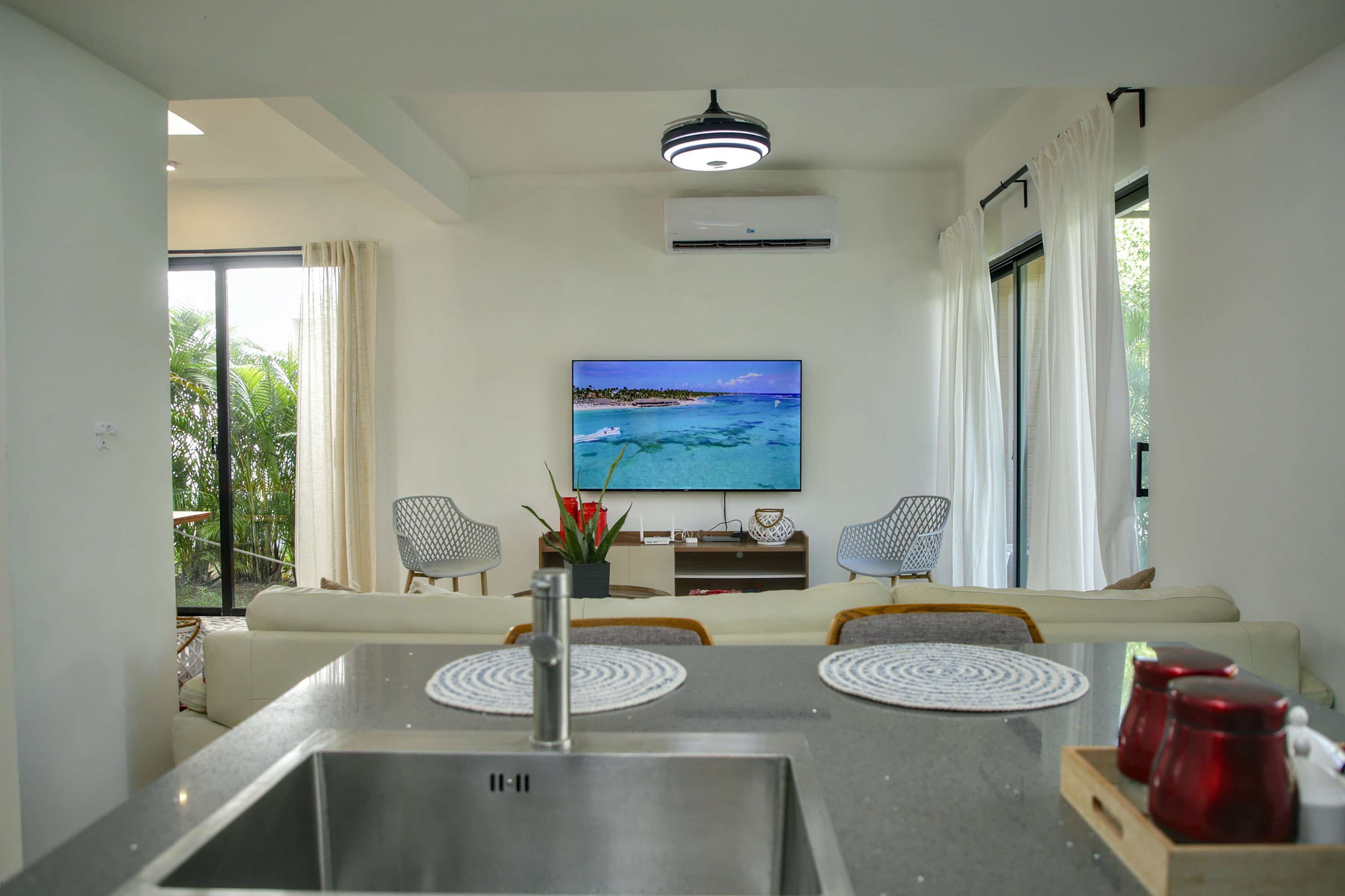 A modern living room with a flat-screen TV, two patterned chairs, a sofa, and a dining area with a stainless steel sink and red mugs.