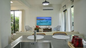 Una sala moderna con un televisor de pantalla plana, dos sillas estampadas, un sofá y una zona de comedor con un fregadero de acero inoxidable y tazas rojas.