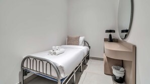 A small, single-bed room with a metal frame, white bedding, a round mirror, a small shelf with a trash bin, and a light-colored wall. - Free Housekeeping Visits During Your Stay - Stayshort - Fresh 2 Bedroom Apartment in JVC That Sleeps 5 (Dubai)