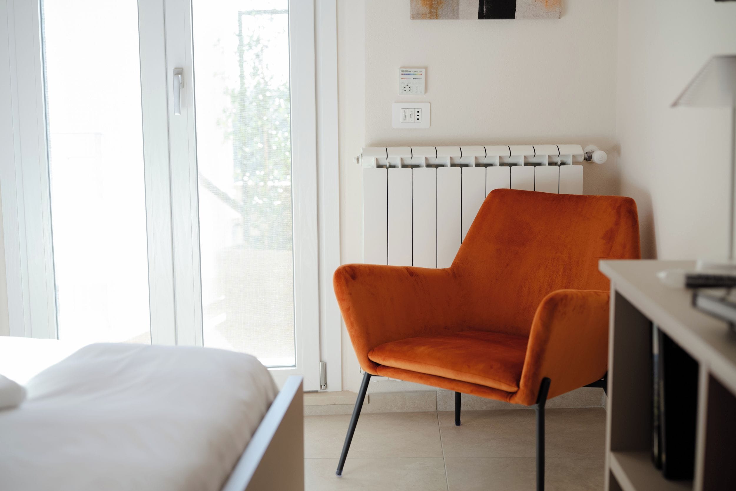 Ein Zimmer mit einem Bett, einem orangefarbenen Sessel, einem Heizkörper und einem Fenster.