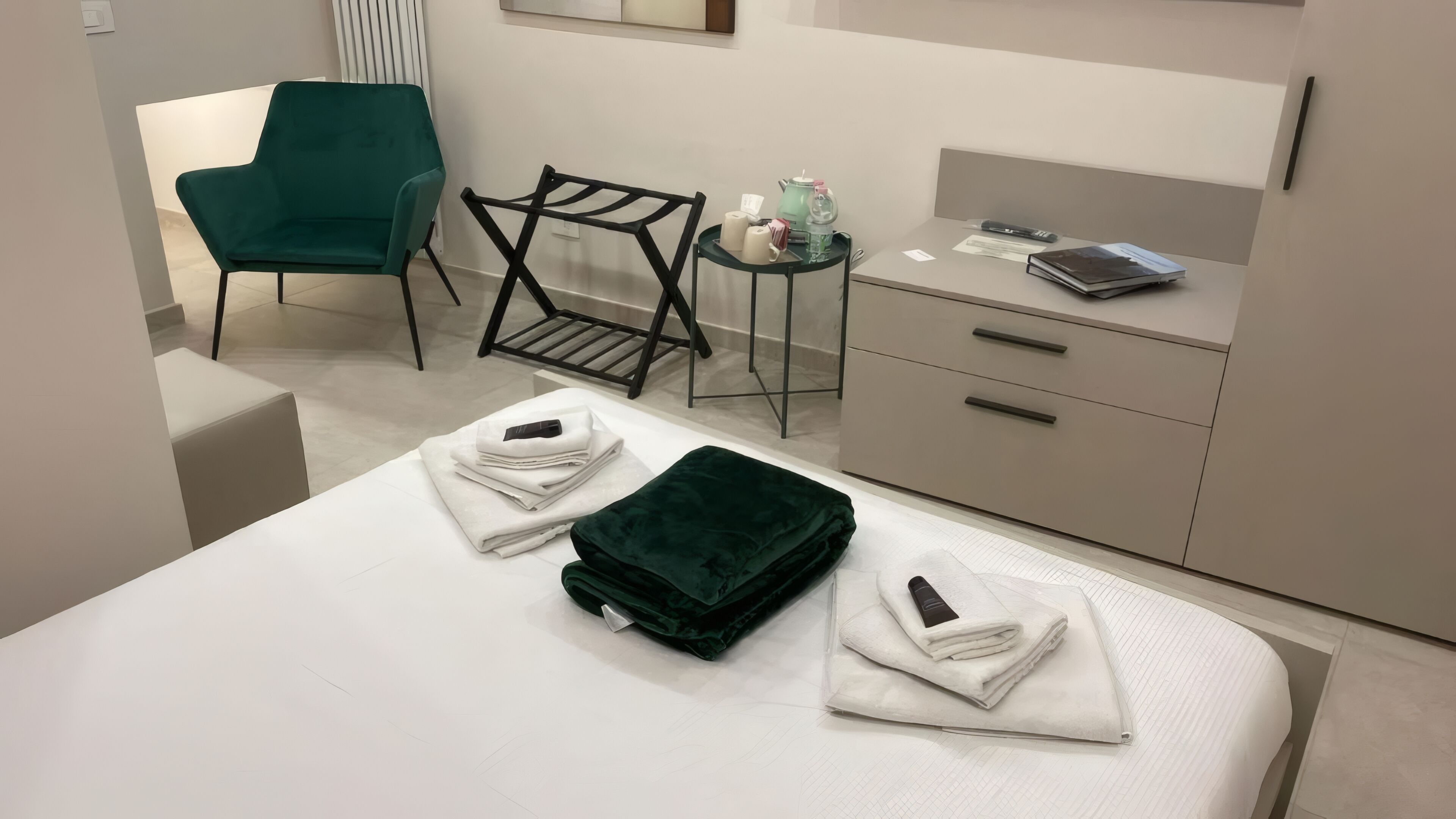 A hotel room with a bed, a green chair, a small table with toiletries, and a desk with a laptop.