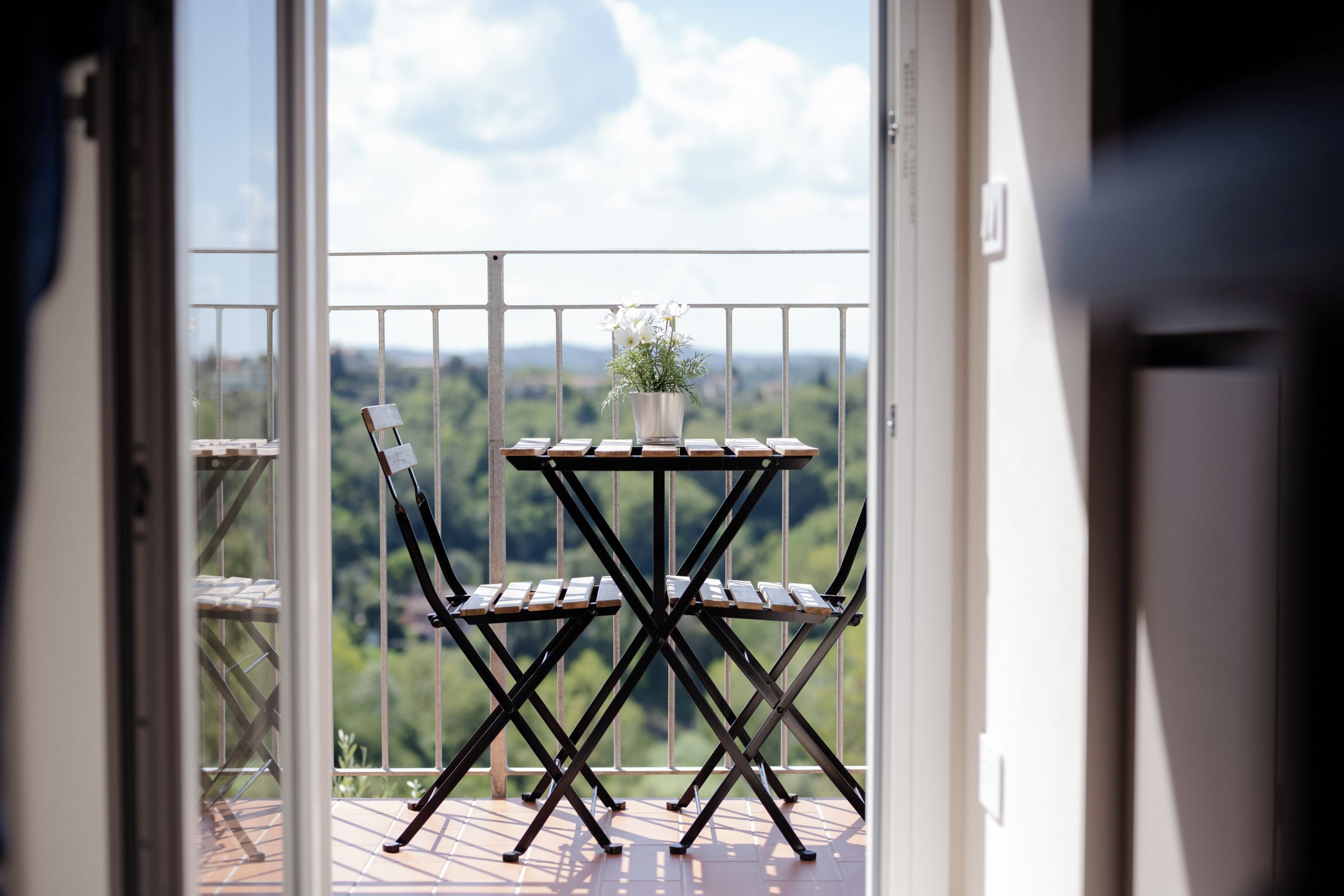 A balcony with a table and chairs, a potted plant, and a view of a green landscape.