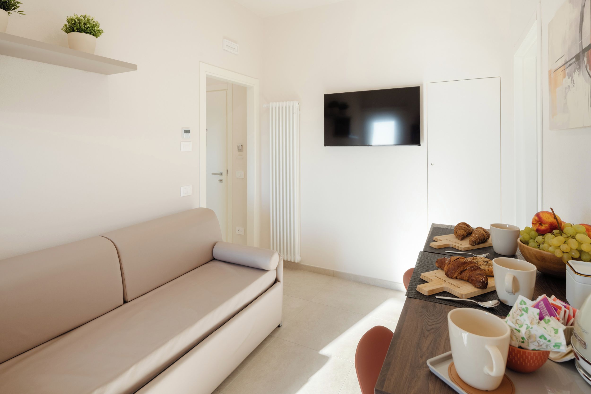 A modern living room with a beige sofa, a dining table with a bowl of fruit and pastries, and a flat-screen TV mounted on the wall.