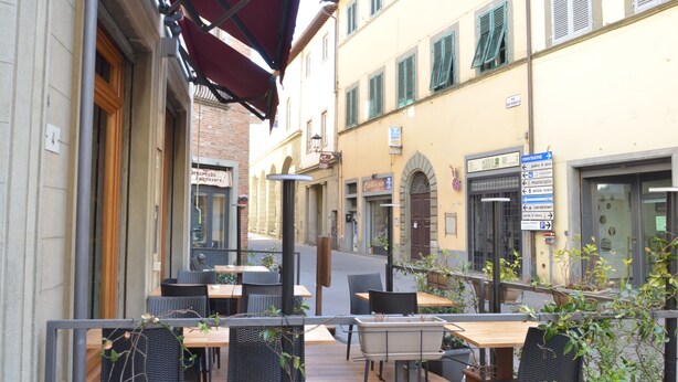Ein Straßencafé mit Tischen und Stühlen, einer roten Markise und Gebäuden mit Schaufenstern im Hintergrund.