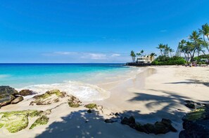 A beach with clear turquoise water, sandy shore, and a building with a pool. - 🏖️Across from Magic Sands | Top Floor Condo w/ AC (Kailua Kona)