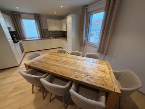 Eine moderne Küche mit weißen Schränken, einem Holztisch mit sechs Stühlen und Blick ins Freie durch ein Fenster.