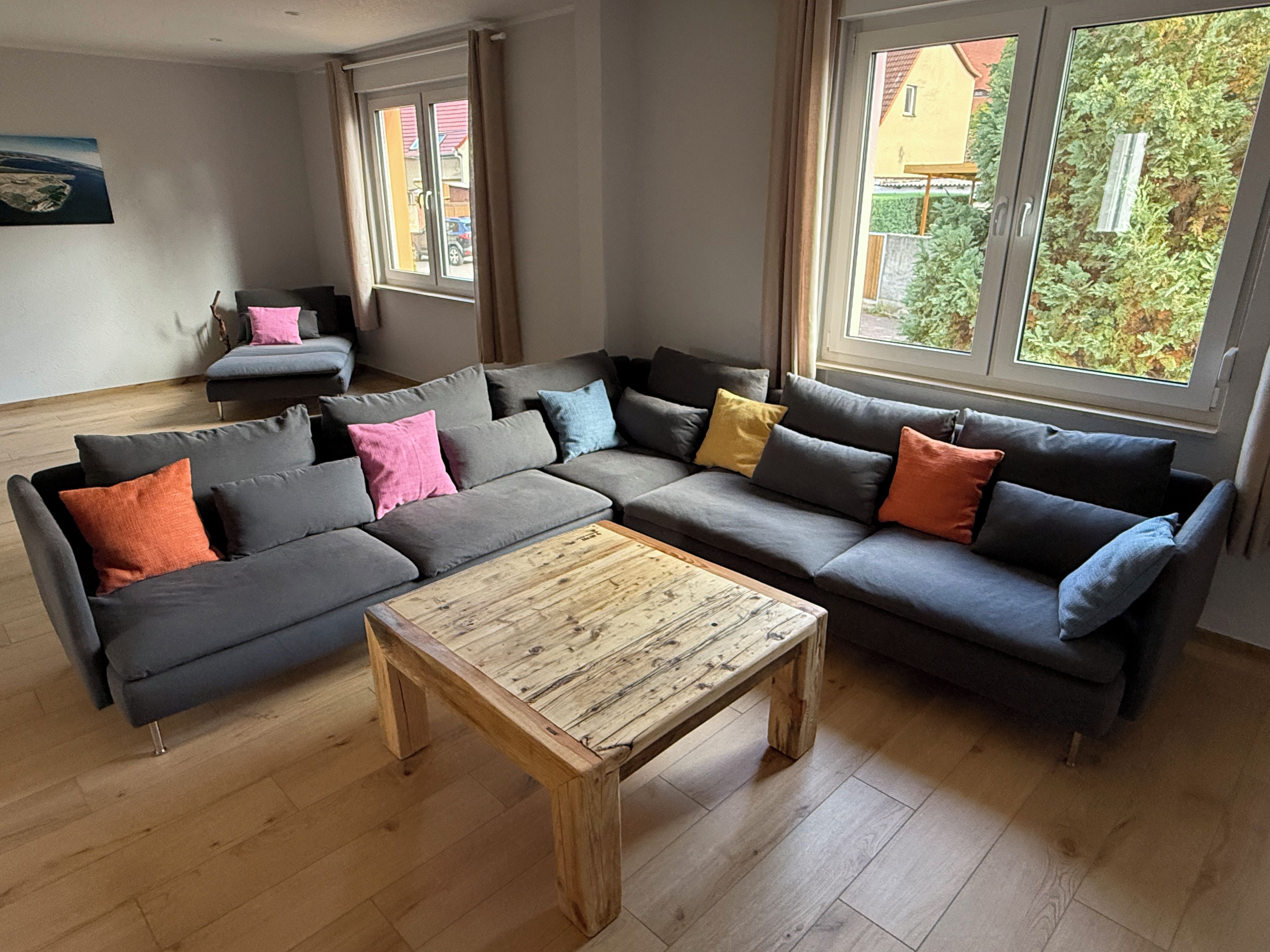 A modern living room with a grey sectional sofa, a wooden coffee table, and large windows offering a view of the outside.
