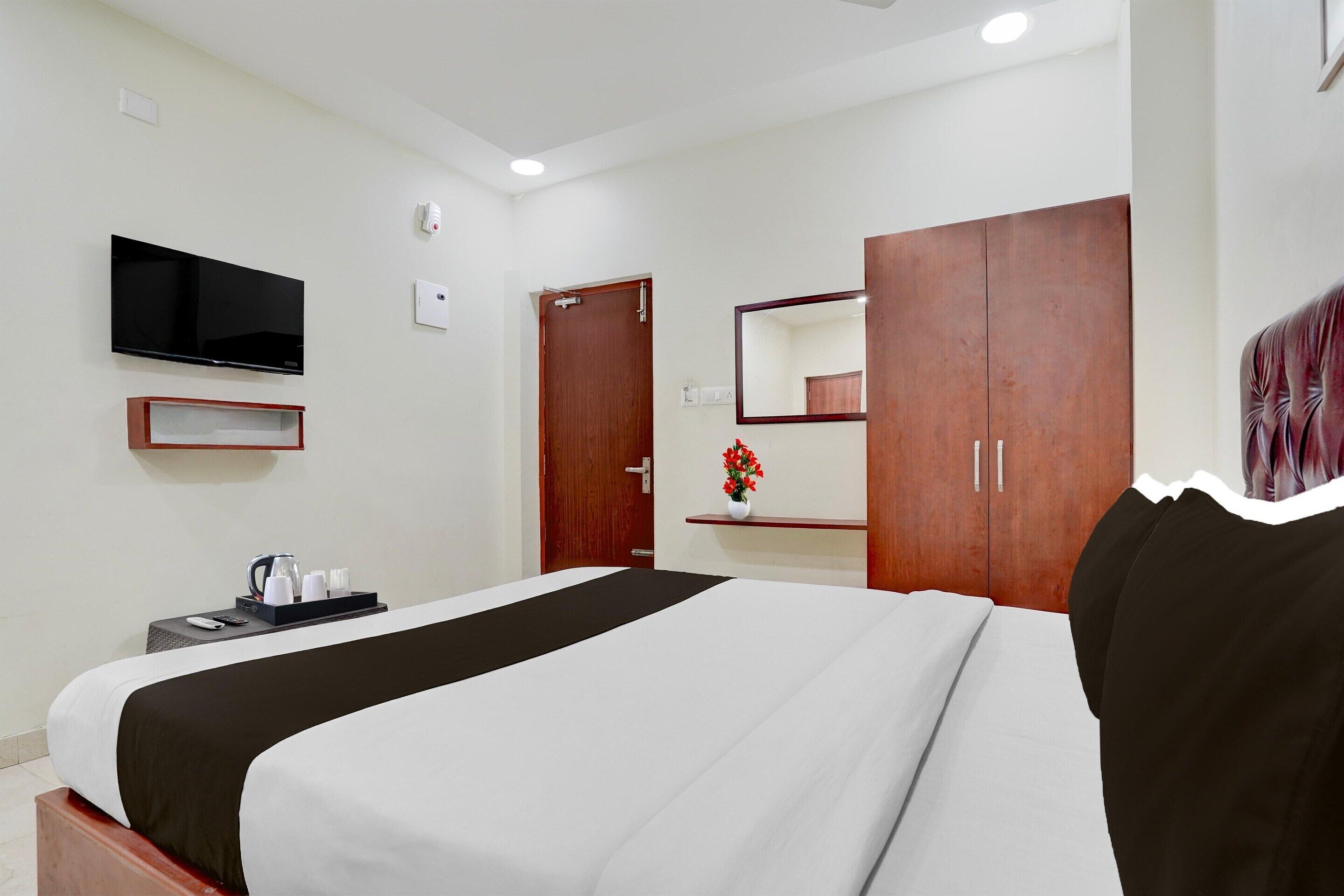 A hotel room with a bed, a TV, a wooden wardrobe, a mirror, and a small shelf with a vase.