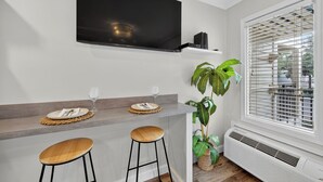 A modern kitchen with a flat-screen TV, two bar stools, a plant, and a window with blinds. - Good Day Sunshine!  Perfect Couples Beach Oasis (Hilton Head Island)